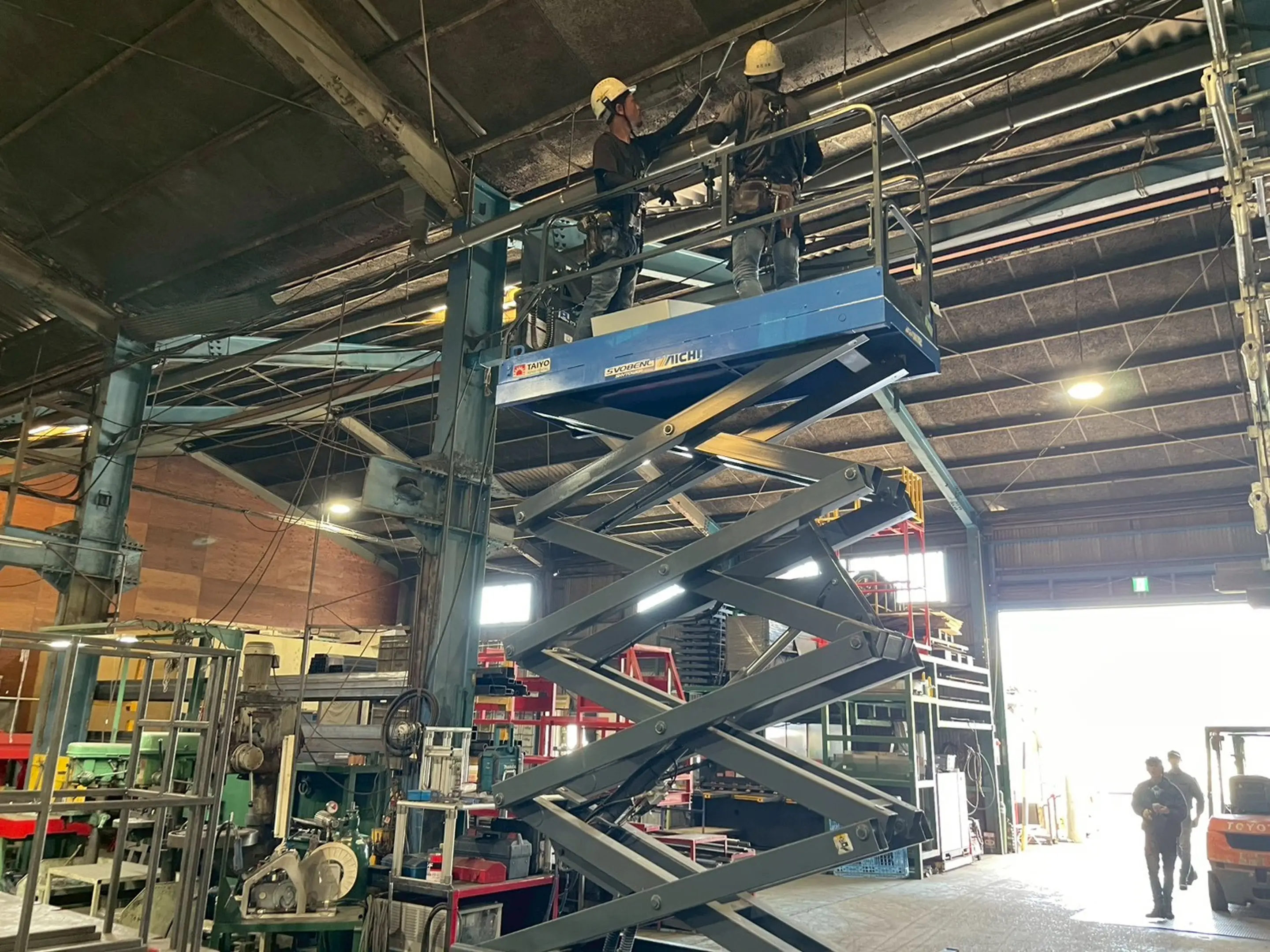 大型機械が並ぶ工場内で昇降式作業台に乗る作業者の全景