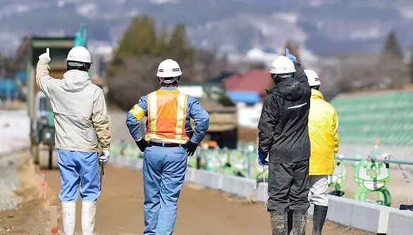 反射ベストや長靴を着用し、整備中の舗装路を背に立つ作業員の後ろ姿